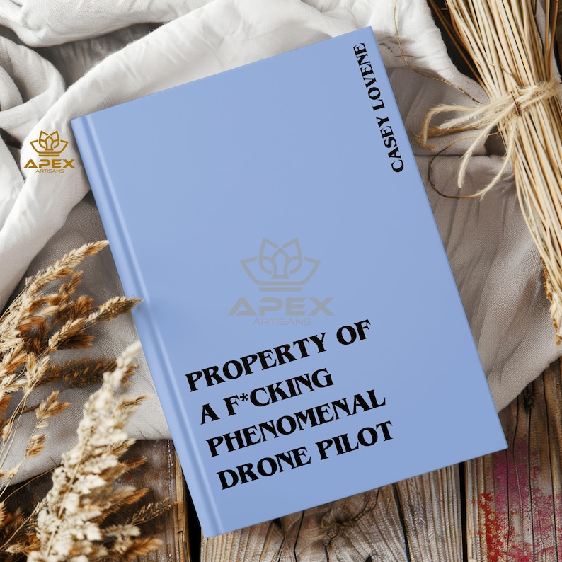 May include: A light blue hardcover book with black text that reads PROPERTY OF A F*CKING PHENOMENAL DRONE PILOT on the front cover and CASEY LOVENE on the spine area. The book features a faint logo in the center and a small logo in the corner.