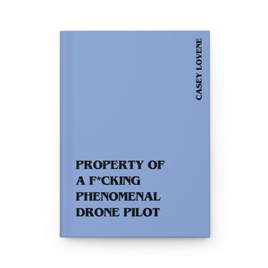 May include: A light blue hardcover journal featuring black text that reads PROPERTY OF A F*CKING PHENOMENAL DRONE PILOT on the front cover, with the name CASEY LOVENE printed vertically in black along the right edge of the front cover.