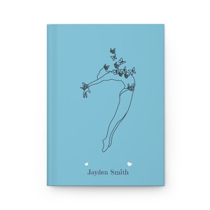 May include: A light blue hardcover journal featuring a black line art illustration of a person leaping with butterflies surrounding them. The name Jayden Smith is printed in dark gray text at the bottom, flanked by two small white heart icons.