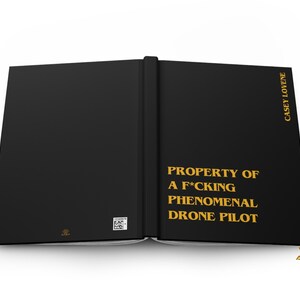 May include: A black hardcover book with gold text that reads PROPERTY OF A F*CKING PHENOMENAL DRONE PILOT on the front cover and CASEY LOVENE on the spine. The back cover features a small square QR code and a tiny gold logo at the bottom.