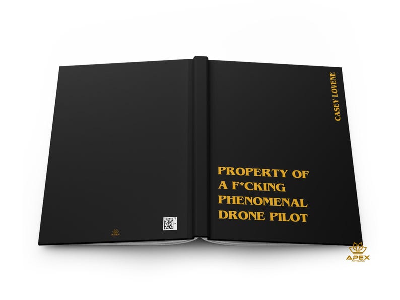 May include: A black hardcover book with gold text that reads PROPERTY OF A F*CKING PHENOMENAL DRONE PILOT on the front cover and CASEY LOVENE on the spine. The back cover features a small square QR code and a tiny gold logo at the bottom.