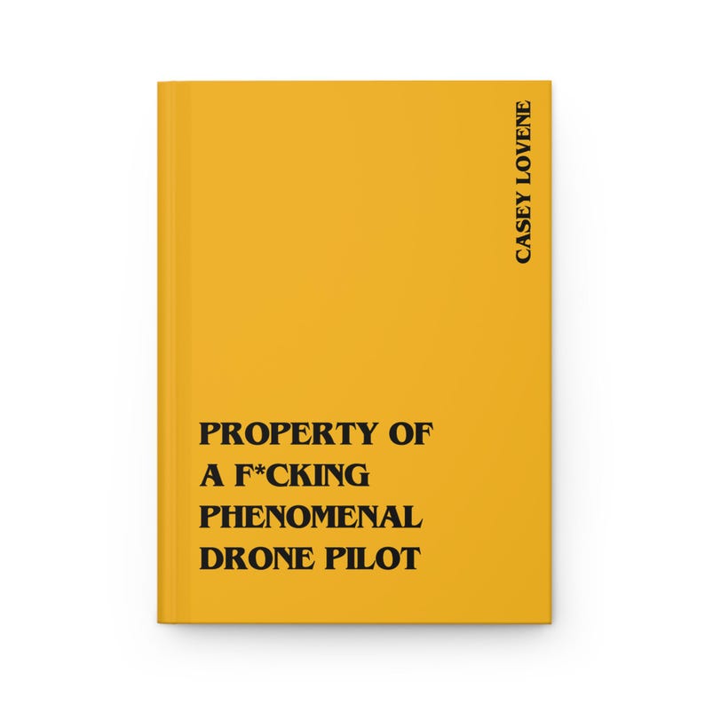 May include: A bright yellow hardcover journal featuring bold black text that reads PROPERTY OF A F*CKING PHENOMENAL DRONE PILOT on the front cover, with the name CASEY LOVENE printed vertically in black along the right edge of the cover.