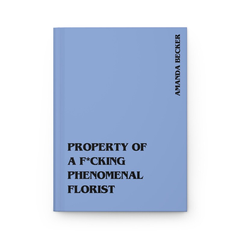May include: A light blue hardcover journal featuring black text that reads PROPERTY OF A F*CKING PHENOMENAL FLORIST on the front cover, with the name AMANDA BECKER printed vertically in black along the right side of the book cover.