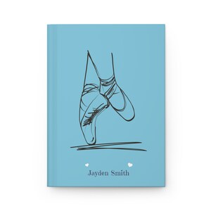 May include: A light blue hardcover journal featuring a black line drawing of ballet slippers on pointe. Below the illustration, the name Jayden Smith is printed in dark gray text, flanked by two small white heart icons on either side of the name.