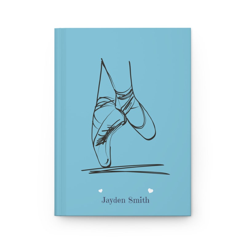 May include: A light blue hardcover journal featuring a black line drawing of ballet slippers on pointe. Below the illustration, the name Jayden Smith is printed in dark gray text, flanked by two small white heart icons on either side of the name.