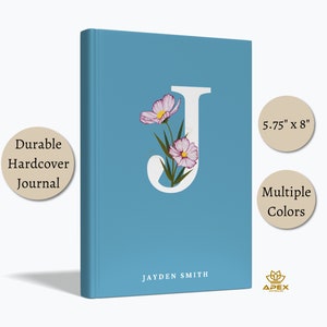 Puede incluir: Un diario de tapa dura azul con una letra J blanca y flores rosas. El diario mide 5,75 pulgadas por 8 pulgadas. El texto "JAYDEN SMITH" está impreso en la portada.