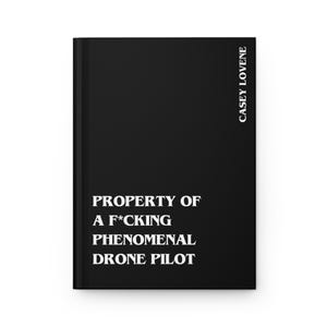 May include: A black hardcover journal with white text on the front cover that reads PROPERTY OF A F*CKING PHENOMENAL DRONE PILOT. The name CASEY LOVENE is printed vertically in white text along the right side of the front cover of the book.