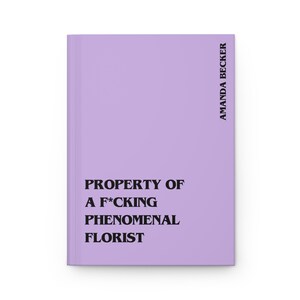 May include: A light purple hardcover journal featuring black text that reads PROPERTY OF A F*CKING PHENOMENAL FLORIST on the front cover, with the name AMANDA BECKER printed vertically in black along the right side of the book cover.