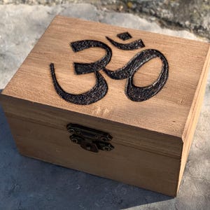 Puede incluir: Una caja de madera con tapa con bisagras y cierre de latón. La parte superior de la caja está decorada con un símbolo Om negro.