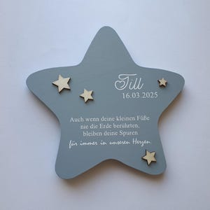 Könnte beinhalten: Ein hellblauer Holzstern mit dem Namen "Till" und dem Datum "16.03.2025" in weiß geschrieben. Der Stern hat vier kleine Holzsterne daran befestigt. Der Text "Auch wenn deine kleinen Füße nie die Erde berührten, bleiben deine Spuren für immer in unseren Herzen." ist in weiß auf dem Stern geschrieben.