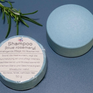 Könnte beinhalten: Zwei hellblaue feste Shampoobars, eine mit einem Etikett mit der Aufschrift "Shampoo [blue rosemary]" und die andere ohne Etikett. Das Etikett enthält Text auf Deutsch und das Gewicht des Produkts beträgt 50 g. Ein Rosmarinzweig ist im Hintergrund.