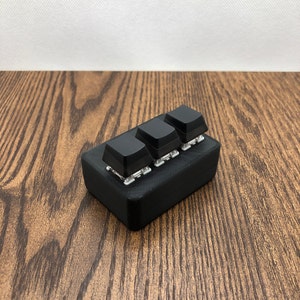 Puede incluir: Un conjunto de tres teclas de teclado negras sobre una base negra. Las teclas cuadradas están sobre un fondo de madera. Las teclas son un componente de un teclado.