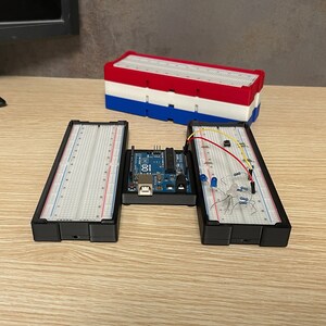 Può includere: Un'impostazione di progetto elettronico con una scheda Arduino blu, breadboard e vari componenti elettronici. Le breadboard sono nere con griglie bianche e linee rosse/blu. Una pila di contenitori di plastica rossi, bianchi e blu è sullo sfondo.