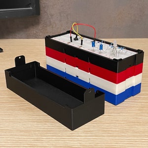 Puede incluir: Una carcasa de plástico negro con un circuito de placa de pruebas. La placa de pruebas tiene varios componentes electrónicos, incluidos LED, resistencias y cables. El circuito está construido sobre una pila de capas de plástico de colores: negro, rojo, blanco y azul.