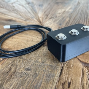 Może przedstawiać: Czarny prostokątny element z trzema srebrnymi przyciskami na górze. Czarny kabel USB jest podłączony do urządzenia i zwinięty na drewnianej powierzchni. Urządzenie wydaje się być panelem sterowania lub przełącznikiem.