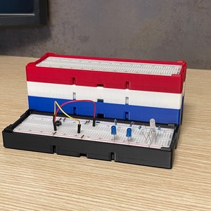 Puede incluir: Una pila de cajas de protoboard rojas, blancas y azules detrás de una protoboard negra abierta. La protoboard contiene componentes electrónicos, incluyendo cables y LEDs, para un proyecto de electrónica DIY.