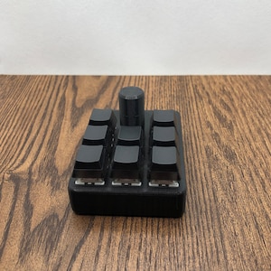 Peut inclure: Un clavier mécanique noir avec neuf touches et un bouton cylindrique. Les touches sont carrées et espacées uniformément, avec une base noire. Le fond est une surface en bois foncé.