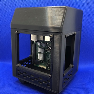 Puede incluir: Una carcasa de ordenador negra impresa en 3D con laterales abiertos, que muestra los componentes internos. La carcasa tiene forma rectangular con una cubierta superior. Los componentes internos incluyen una placa de circuito verde.