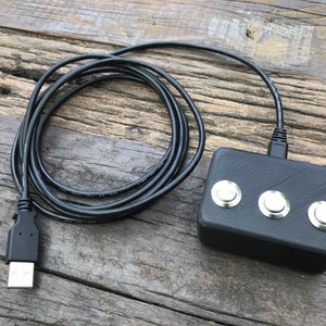 Peut inclure: Un appareil électronique noir avec trois boutons argentés, connecté à un câble USB noir. L'appareil a une surface texturée et est posé sur un fond en bois vieilli.