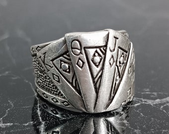 Puede incluir: Anillo plateado con diseños de naipes. El anillo muestra la Reina y la Jota de diamantes, con detalles intrincados en la banda. El metal tiene un aspecto ligeramente envejecido.
