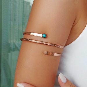 Può includere: Bracciale da braccio color rame con un accento di pietra turchese. La fascia superiore ha un design piatto, mentre la fascia inferiore presenta un motivo inciso. Il gioiello avvolge il braccio, un accessorio unico ed elegante.
