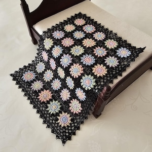 Könnte beinhalten: Eine schwarze Häkeldecke mit einem Blumenmuster in Pastellfarben, drapiert über einem Miniaturbett. Die Decke hat ein sich wiederholendes Design aus bunten Blumen mit einem gewellten Rand. Der Bettrahmen ist dunkelbraun.
