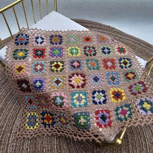 May include: A handmade crochet blanket featuring a grid of multicolored granny squares joined by light brown yarn. Each square displays a unique combination of vibrant colors including red, blue, yellow, green, and purple, finished with a delicate lace border.