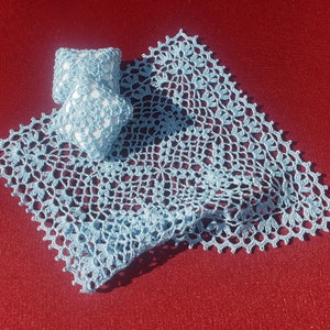 Może przedstawiać: Ażurowa serwetka w jasnoniebieskim kolorze z delikatnym wzorem kwiatowym. Serwetka leży na czerwonym tle i ma na sobie dwa małe białe kwadraty wykonane na szydełku.