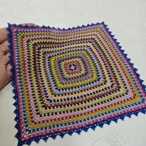 May include: A square crochet doily featuring a vibrant, multi-colored granny square pattern with concentric rows of various hues. The piece is finished with a decorative, pointed dark blue crochet border along all four edges of the square.