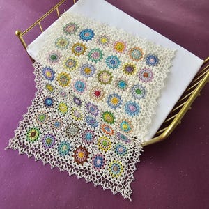 May include: A handmade, off-white crochet blanket with colorful floral motifs. The blanket features a square pattern with a scalloped edge, showcasing a variety of colors including blue, yellow, green, and pink. The blanket is draped over a white surface.