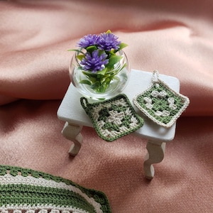 Może przedstawiać: Dwa zielono-białe szydełkowe uchwyty na garnki na białym stole z szklanym wazonem z fioletowymi kwiatami.