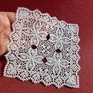 Könnte beinhalten: Eine weiße Häkeldeckchen mit quadratischer Form und filigranen Blumendesigns.