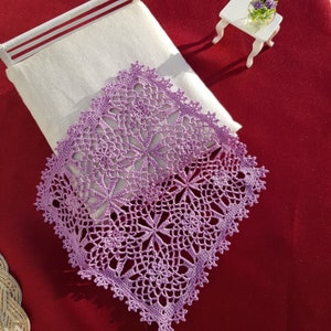 May include: A square, lavender-colored crocheted doily with a floral pattern, resting on a white pillow. The doily has a detailed, openwork design and a scalloped edge. The background is a deep red surface.