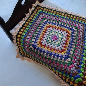 May include: A colorful, handmade crochet blanket with a square pattern. The blanket features a variety of colors, including pink, blue, green, yellow, and brown. It is placed on a white surface and has a decorative, scalloped edge.