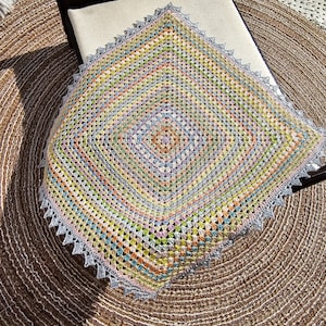 May include: A handmade, square crochet blanket in a granny square pattern. The blanket features a vibrant mix of pastel colors, including pink, yellow, blue, and green. The edges are finished with a light blue, scalloped trim. The blanket is approximately 36 inches square.