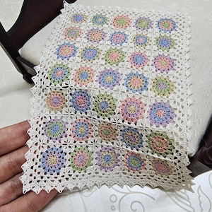 May include: A handmade, square crochet doily featuring a repeating floral pattern in pastel colors. The doily is primarily white with delicate, colorful flowers in pink, blue, yellow, and green. The edges are finished with a scalloped trim.