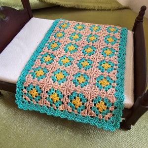 May include: A handmade crochet blanket in a floral pattern, featuring a color palette of peach, teal, and yellow. The blanket is placed on a small wooden bed. The edges are finished with a teal scalloped border.