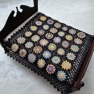 May include: A black crocheted blanket with colorful floral designs. The blanket is draped over a small, dark wooden bed frame. The flowers are in shades of yellow, pink, blue, green, and purple.