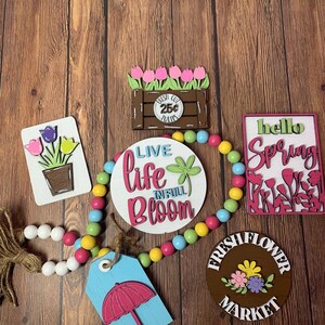 May include: A collection of spring-themed wooden signs with a variety of colors and designs. The signs include "Live Life in Full Bloom", "Fresh Flower Market", "Hello Spring", and a sign with a flower pot and tulips. A beaded garland with white, yellow, pink, and blue beads is also included.