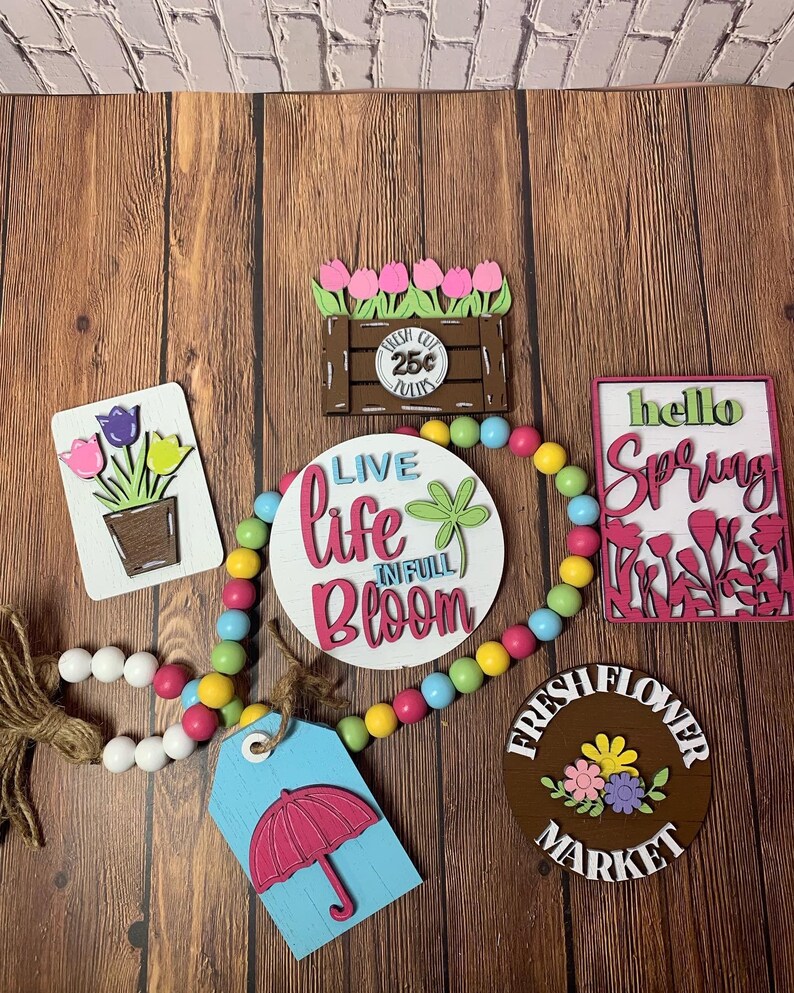 May include: A collection of spring-themed wooden signs with a variety of colors and designs. The signs include "Live Life in Full Bloom", "Fresh Flower Market", "Hello Spring", and a sign with a flower pot and tulips. A beaded garland with white, yellow, pink, and blue beads is also included.