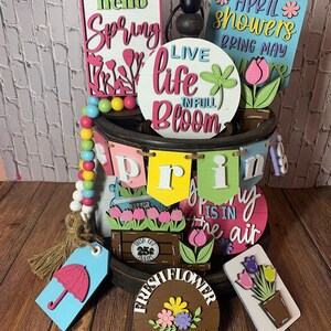 May include: A wooden tiered tray decorated with spring-themed signs and decorations. The signs feature phrases like "hello spring", "live life in full bloom", and "April showers bring May flowers". There are also wooden tulips, a wooden umbrella, and a wooden sign that says "fresh flower market".