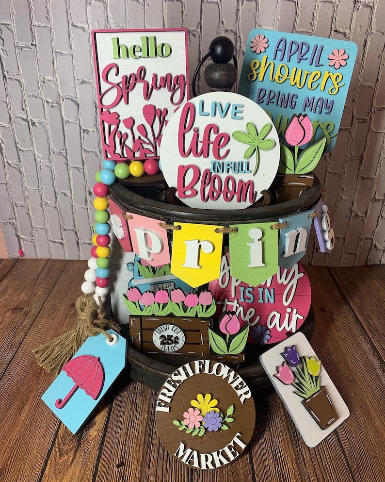 May include: A wooden tiered tray decorated with spring-themed signs and decorations. The signs feature phrases like "hello spring", "live life in full bloom", and "April showers bring May flowers". There are also wooden tulips, a wooden umbrella, and a wooden sign that says "fresh flower market".