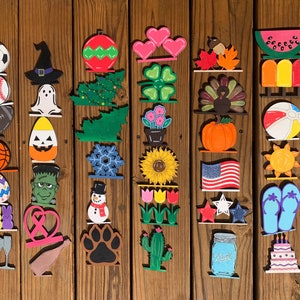 May include: A collection of 20 wooden painted ornaments featuring various themes, including sports, holidays, and nature. The ornaments are arranged in rows on a wooden surface.