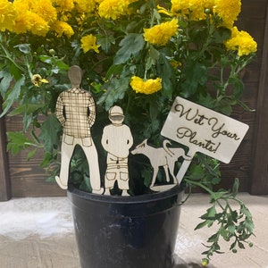 May include: A black plastic pot with a yellow flower plant. The plant has a wooden sign that says "Wet Your Plants!" and wooden figures of a man, a boy, and a dog.
