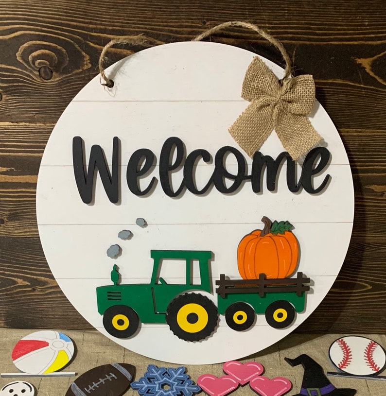 May include: A round wooden welcome sign with a white background and black lettering. The sign features a green tractor pulling a wagon with a large orange pumpkin. A burlap bow is attached to the top of the sign.