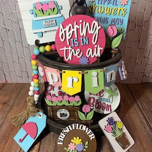 May include: A tiered tray decorated with spring-themed wooden signs. The signs feature phrases like "Spring is in the air", "April showers", and "Fresh Flower Market". There are also wooden cutouts of tulips, a truck, and a potted plant.