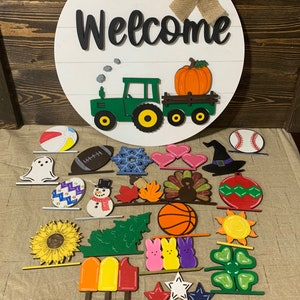 May include: A round wooden sign with the word "Welcome" painted in black. A green tractor with a yellow pumpkin in the back is painted on the sign. There are 24 different wooden shapes painted in various colors and designs, including a ghost, a snowman, a turkey, a basketball, a sunflower, a popsicle, a watermelon, and a four-leaf clover.