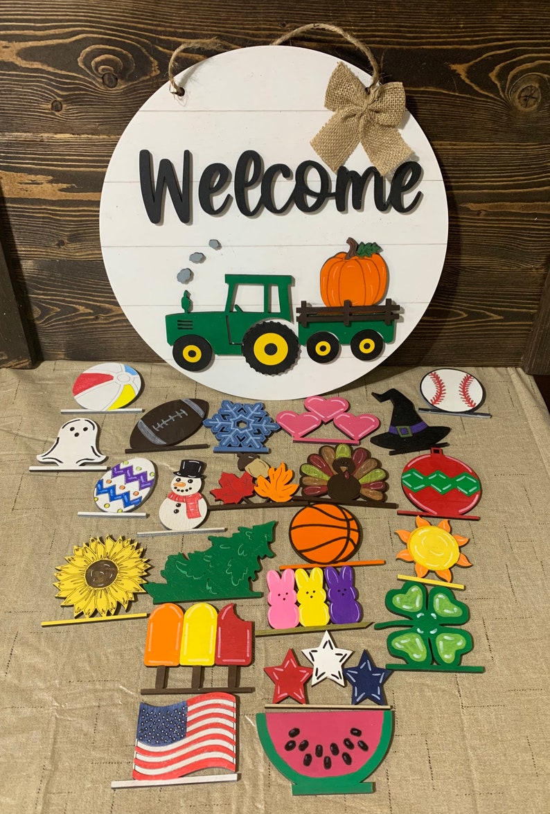 May include: A round wooden sign with the word "Welcome" painted in black. A green tractor with a yellow pumpkin in the back is painted on the sign. There are 24 different wooden shapes painted in various colors and designs, including a ghost, a snowman, a turkey, a basketball, a sunflower, a popsicle, a watermelon, and a four-leaf clover.