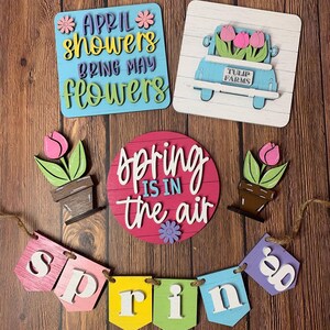 May include: A set of spring-themed wooden signs with a banner that reads "spring". The signs feature flowers, a truck with flowers, and the phrase "April showers bring May flowers".