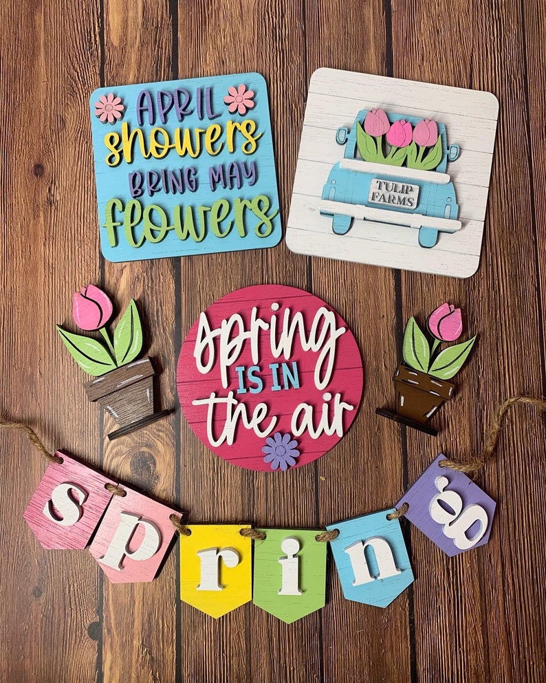 May include: A set of spring-themed wooden signs with a banner that reads "spring". The signs feature flowers, a truck with flowers, and the phrase "April showers bring May flowers".